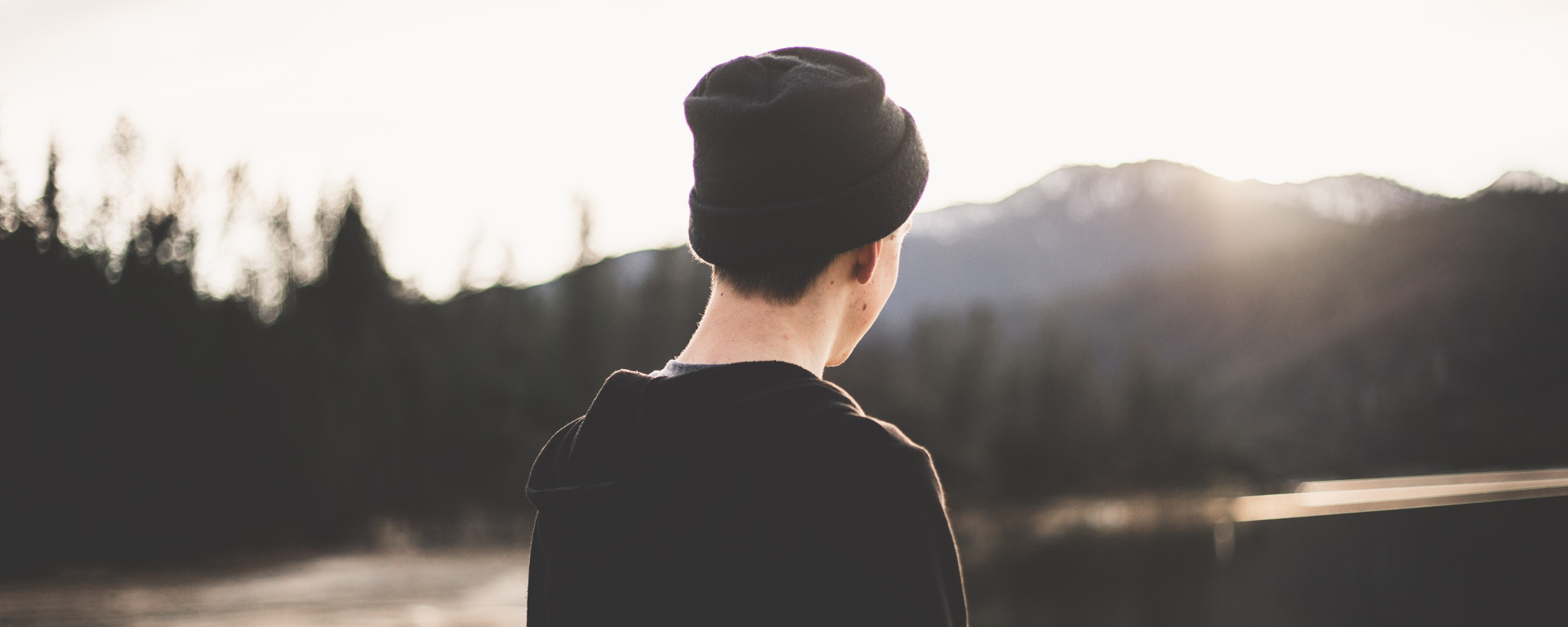 A boy with a black beanie watching a river landscape. The sun shines on his face from behind the mountains in the background.