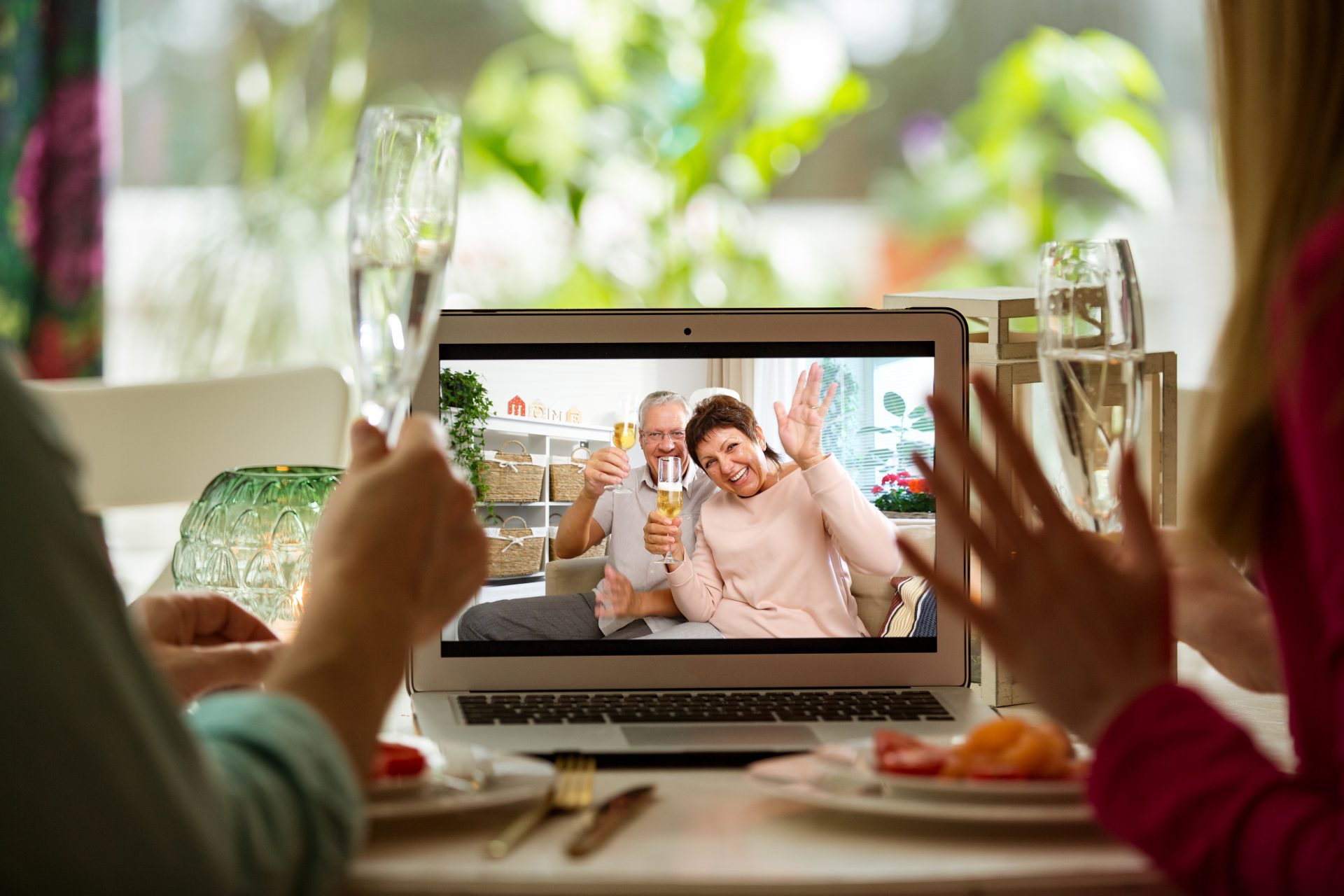 A man and a woman on a video call with older parents. They are eating and toasting with sparkling wine..
