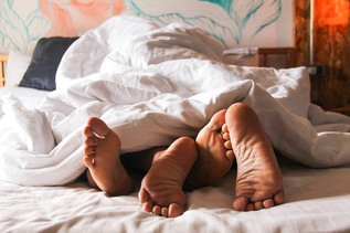 Sängyssä kahden ihmisen jalat, jotka pilkistävät peiton alta.