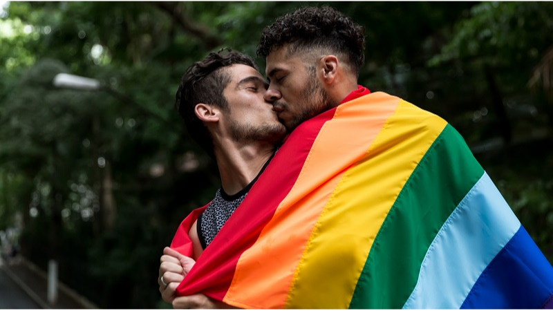 kaksi miestä suutelee kääriytyneenä sateenkaarilippuun