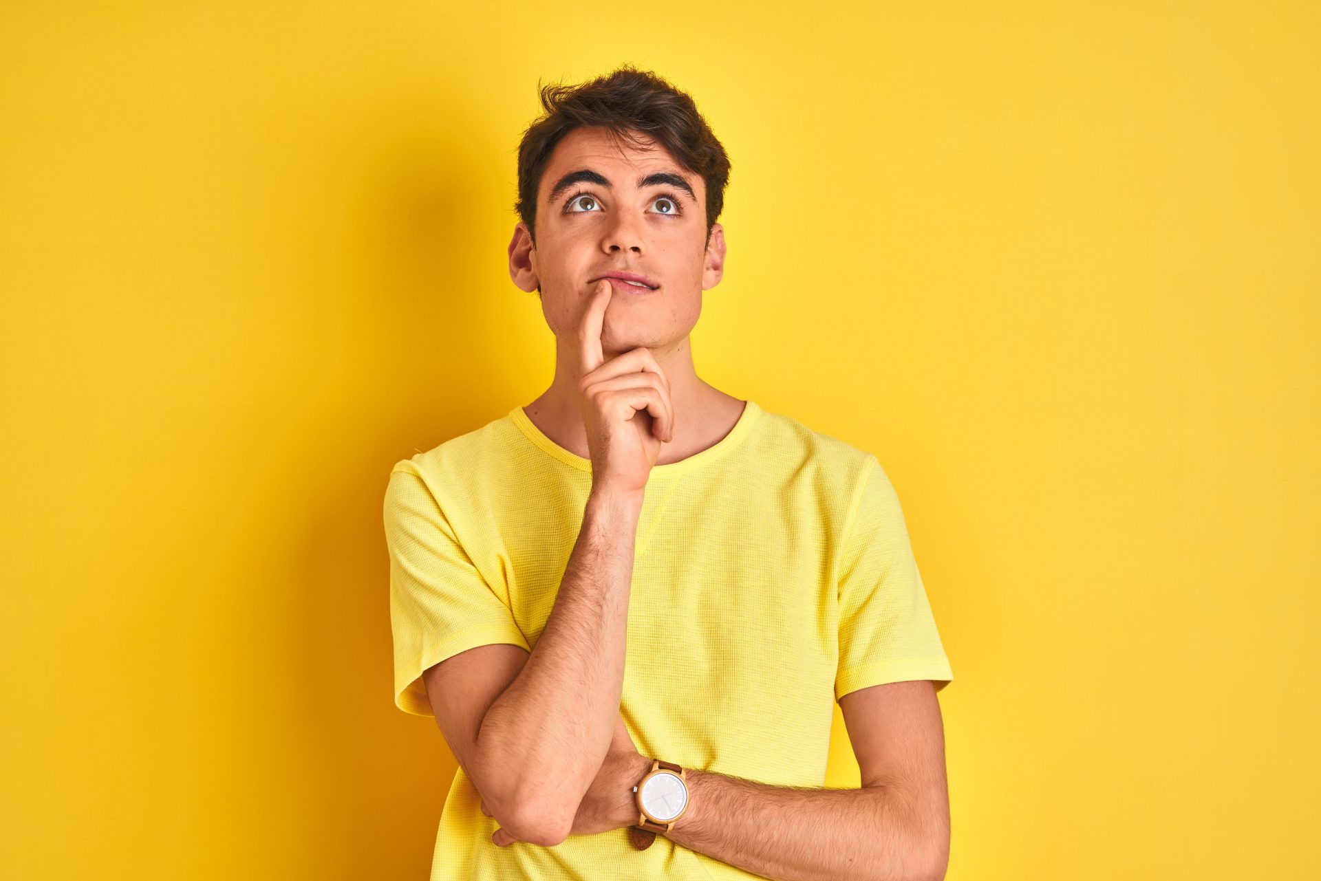 A boy standing with his back against a yellow wall, wearing a yellow T-shirt. He is holding his right hand under his cheek, watching upwards, looking thoughtful.