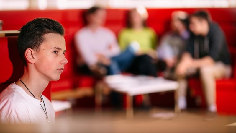 Pojan siluetti, hän katsoo etäisyyteen ja taustalla näkyy hämärästi ryhmä muita nuoria.