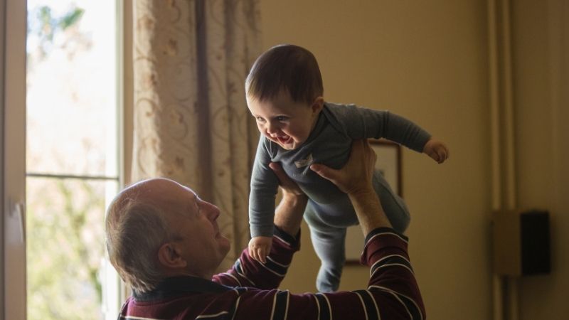 Isoisä pitää pientä lasta ojennetuin käsivarsin ylhäällä ja lapsi nauraa.