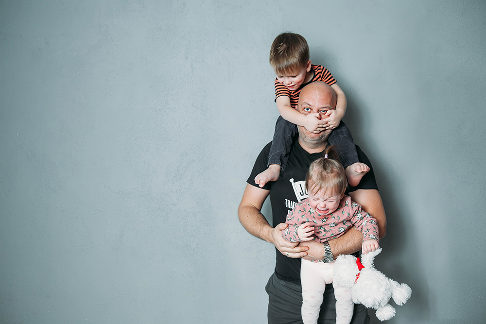 Isällä on reppuselässä yksi lapsi ja toinen on sylissä. Asento on hankala ja tilanne hauska mutta myös stressaava.. stressed young dad with two small children in his arms.