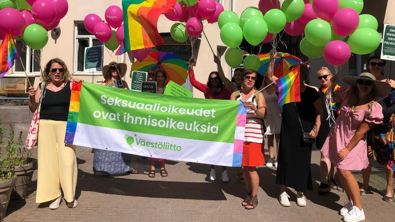 Väestöliittolaiset kokoontuvat Helsinki Pride-kulkueeseen 2022 Väestöliiton sisäpihalla. He pitävät käsissään sateenkaarilippuja, ilmapalloja ja yhteistä banderollia tekstillä "Seksuaalioikeudet ovat ihmisoikeuksia".