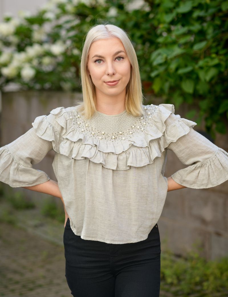 Vaaleaihoinen henkilö, jolla vaaleat hiukset. Päällä harmaanvalkoinen paita jossa röyhelöä rintakehällä ja olkapäillä.