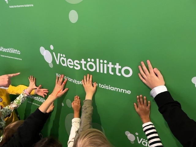 Lasten käsiä Väestöliiton logoilla koristellut vihreän taustan päällä.